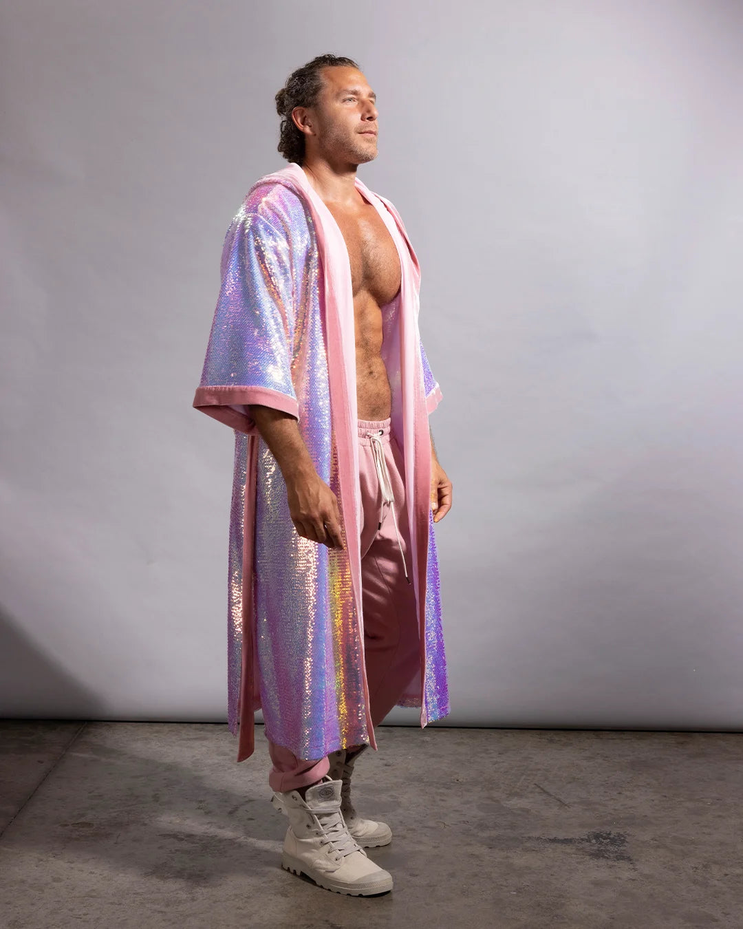 A man stands in profile wearing a sparkling iridescent sequin festival kimono over a matching pink outfit and white boots, photographed against a plain light gray studio backdrop.