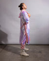 A person standing in profile wearing a long iridescent pink and purple hooded robe or cover-up over pink pants and off-white combat boots, photographed against a plain light gray studio backdrop.