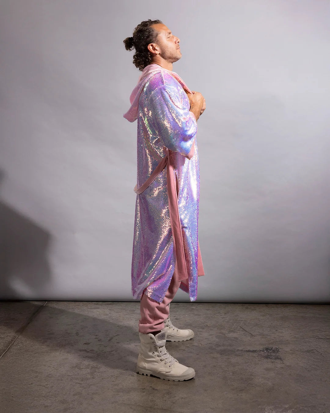 A person standing in profile wearing a long iridescent pink and purple hooded robe or cover-up over pink pants and off-white combat boots, photographed against a plain light gray studio backdrop.