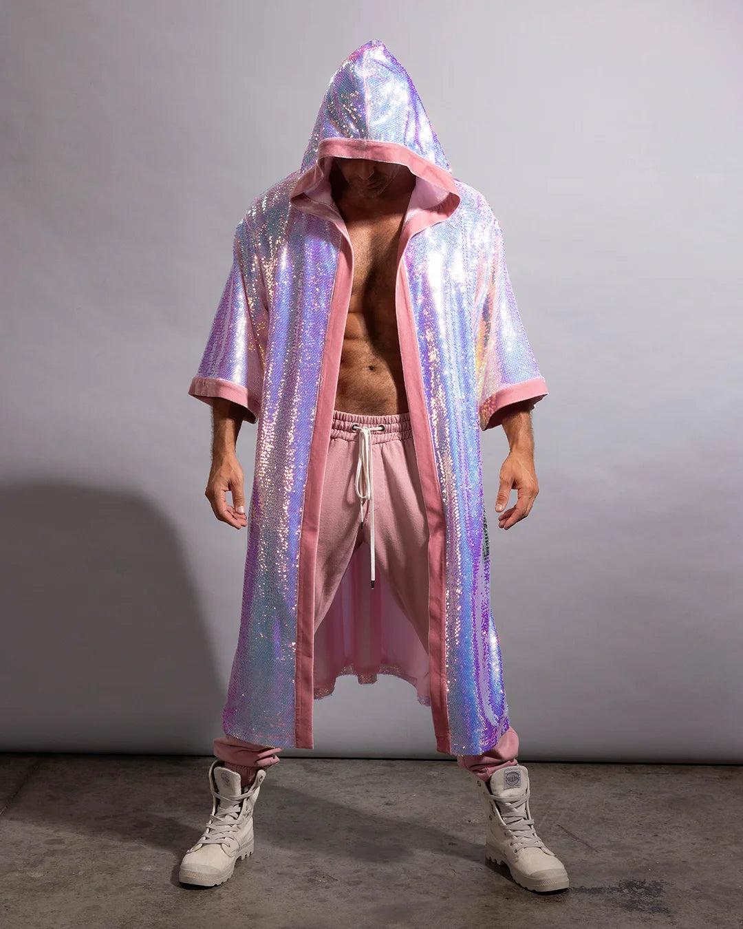 A person modeling a long iridescent holographic hooded kimono robe with pink trim, worn open over pink drawstring pants and white high-top sneakers, against a plain gray studio backdrop.