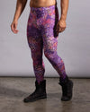 A man modeling high-waisted leggings with a purple, pink, and orange leopard-like abstract pattern, paired with black boots against a gray studio backdrop.