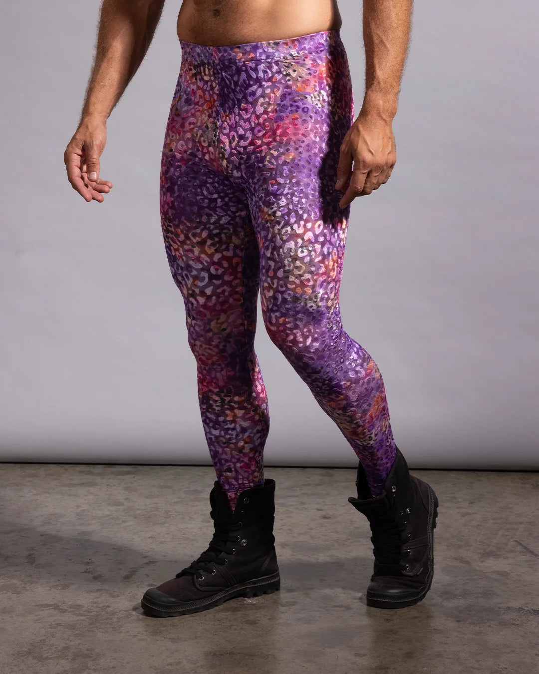 A man modeling high-waisted leggings with a purple, pink, and orange leopard-like abstract pattern, paired with black boots against a gray studio backdrop.