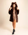 A Woman walking towards the camera, smiling, wearing a plush luxury faux fur coat in deep chocolate brown, with an oversized hood and wolf ears on top, she is holding the plush ultra-soft hood with the coat being worn open. SpiritHoods Custom Made Fake Fur Jackets and Animal Print Coats.