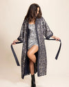 Woman wearing a long, Kimono-Robe with silver and gold sequins in a leopard print pattern over a sequined outfit on a beige background
