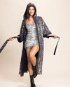 Woman wearing a deep blue denim light weight kimono stitched with silver and gold sparkling sequins into a eyecatching leopard print pattern, over a white dress against a beige background