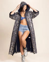 Woman wearing a long leopard-patterned kimono with hood and belt-tie, featuring eye-catching silver and gold sequins creating the animal print pattern throughout the Hooded Kimono-Robe, over a blue top and denim shorts on a beige background