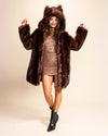 A Woman with her hands up dancing towards the camera, smiling, wearing SpiritHoods plush luxury brown wolf inspired fake fur coat with a hood and wolf ears on a beige background. SpiritHoods Made to Order Faux Fur Coats and Jackets