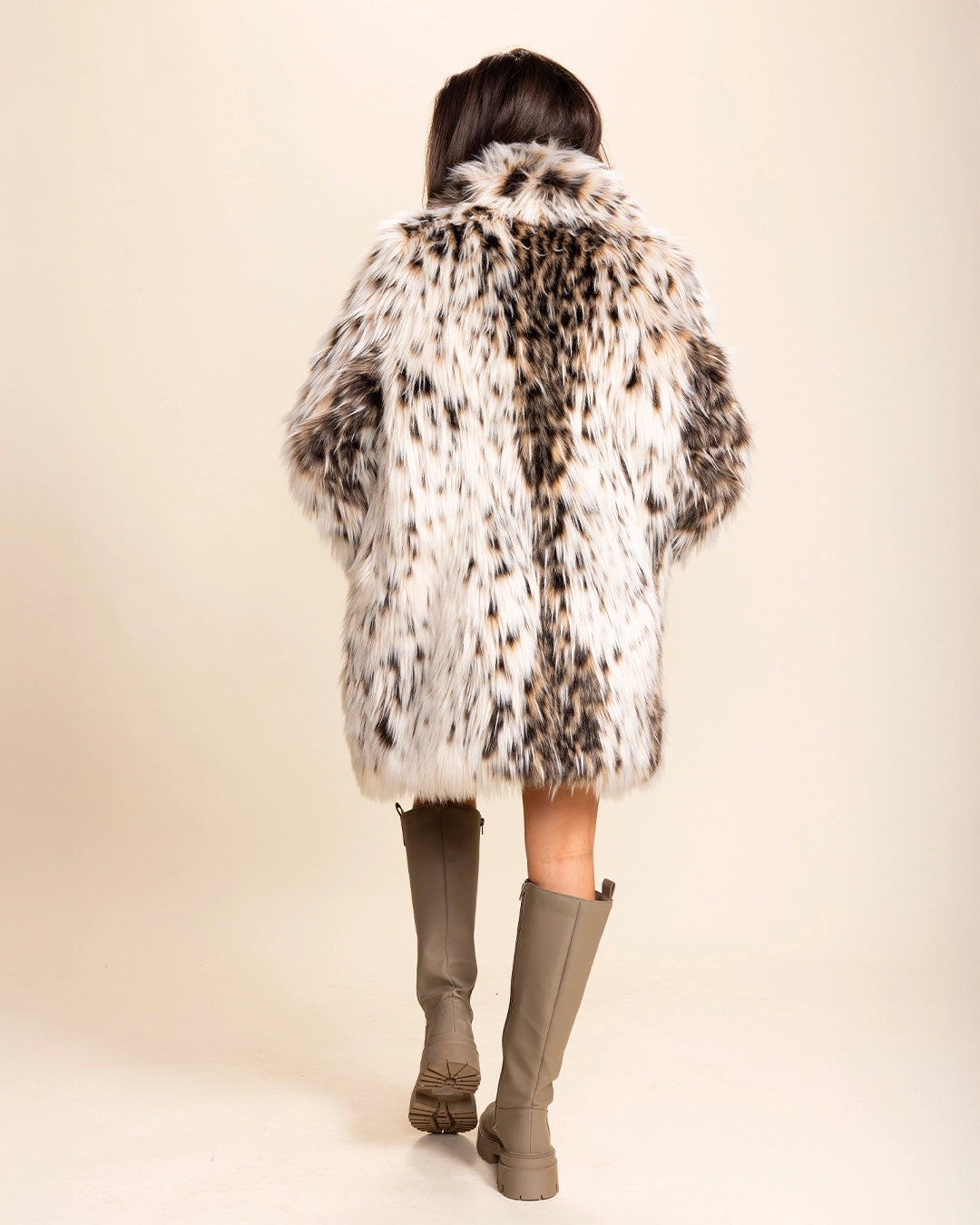 A Woman facing away from the camera wearing a cheetah pattern fake fur jacket in white and brown hues, and knee-high boots on a beige background. Spirit Hoods Custom Made Faux Fur Coats and Plush Luxury Animal Print Jackets