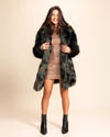 Woman wearing a thigh length, dark green and brown faux fur coat over a sparkly dress on a beige background. SpiritHoods Custom Made Fake Fur Animal inspired Coats and Jackets.