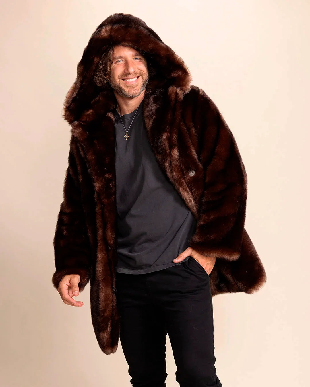 Man wearing a brown fake fur coat against a beige background, smiling wearing a vegan faux fur jacket with hood and peacoat buttons.