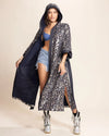 Woman wearing a long shimmering sequin leopard print kimono with a hood, over denim shorts, and silver boots on a beige background