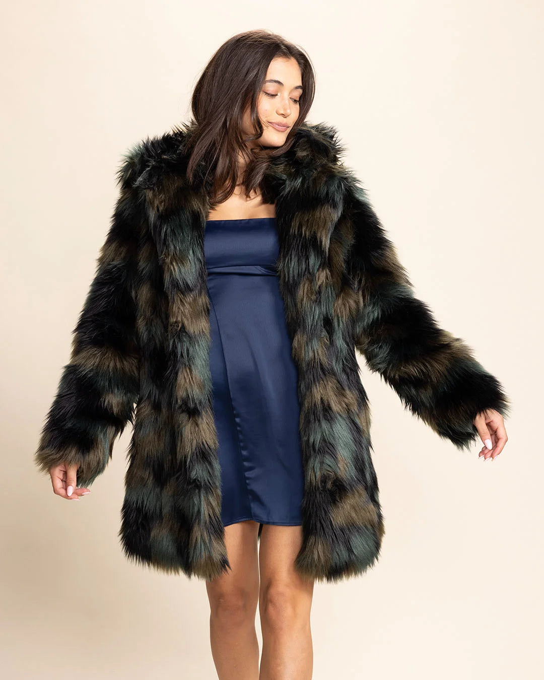 Woman wearing a forest green, brown, and black fake fur coat with arms outstretched on a beige background