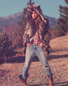 A Woman in nature wearing SpiritHoods classic leopard print fake fur jacket with the hood up showing leopard ears on the top, while walking in the mountains surrounded by trees.