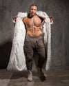 Man with a white fake fur floor length coat and minimal clothing against a textured gray wall.