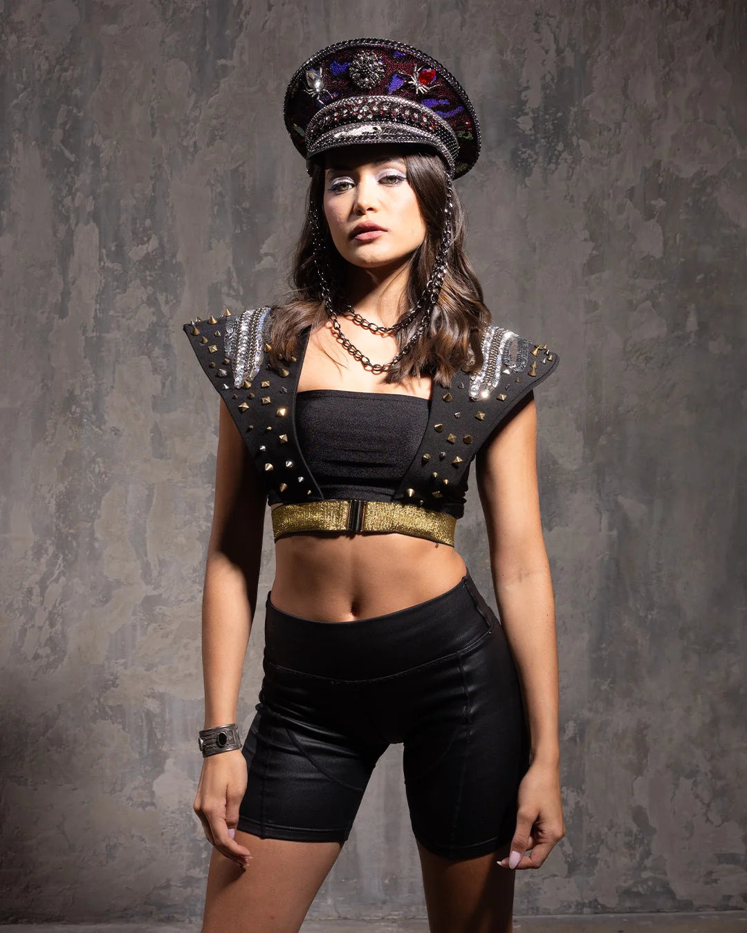 Woman wearing a Festival Captains Hat with vibrant red and purple sequins, and gemstones, wearing a black outfit with decorative elements against a textured gray background