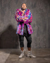 Man wearing a colorful blue and pink neon leopard print coat with his arms crossed against a textured gray wall