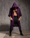 A Man wearing a long purple hooded leopard print kimono against a textured gray wall.