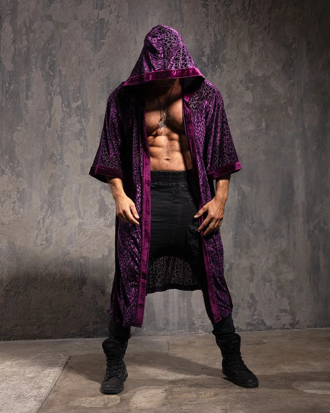 A Man wearing a long purple hooded leopard print kimono against a textured gray wall.