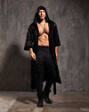 Man wearing a black sequin hooded kimono with a sparkling snakeskin pattern over dark pants against a textured gray wall.