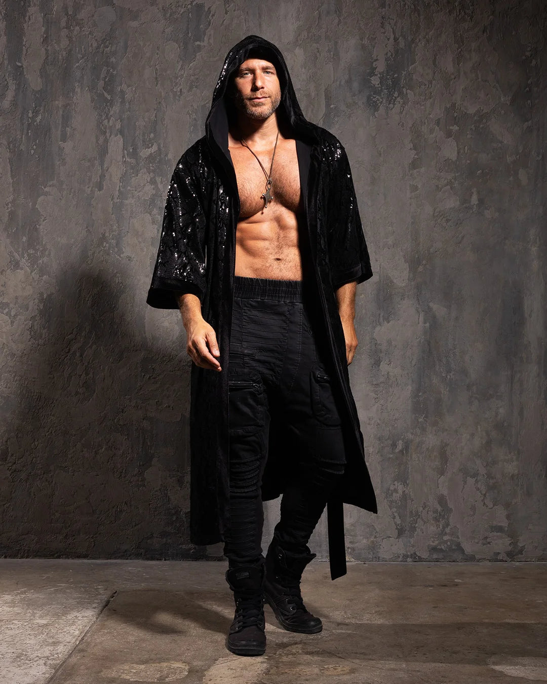 Man wearing a black sequin hooded kimono with a sparkling snakeskin pattern over dark pants against a textured gray wall.