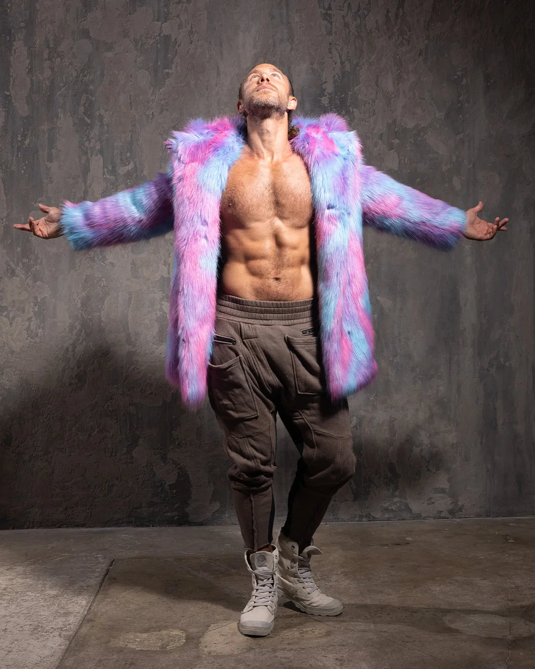 Man wearing wearing a colorful tie dye pink and blue faux fur coat with oversized hood and bear ears, against a textured wall.