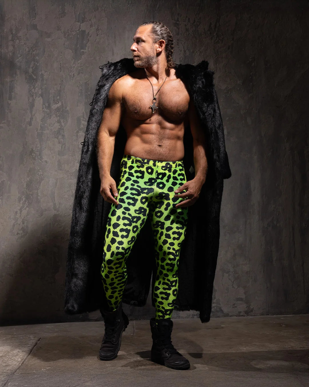 Man wearing neon green leopard print velvet leggings with a black faux fur coat over his shoulder against a gray background
