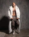 Man wearing a long white leopard print faux fur coat over a brown outfit against a textured gray wall.
