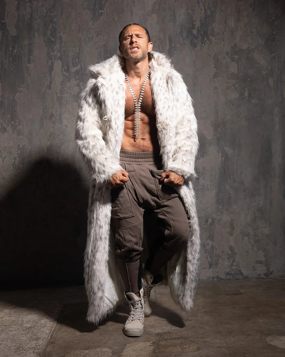 Man wearing a long white leopard print faux fur coat over a brown outfit against a textured gray wall.
