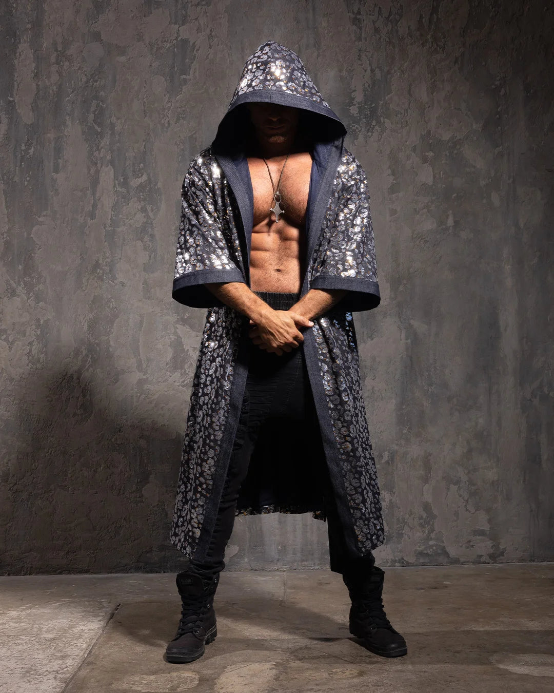 Man wearing a silver and gold leopard print sequined hooded kimono against a textured wall.