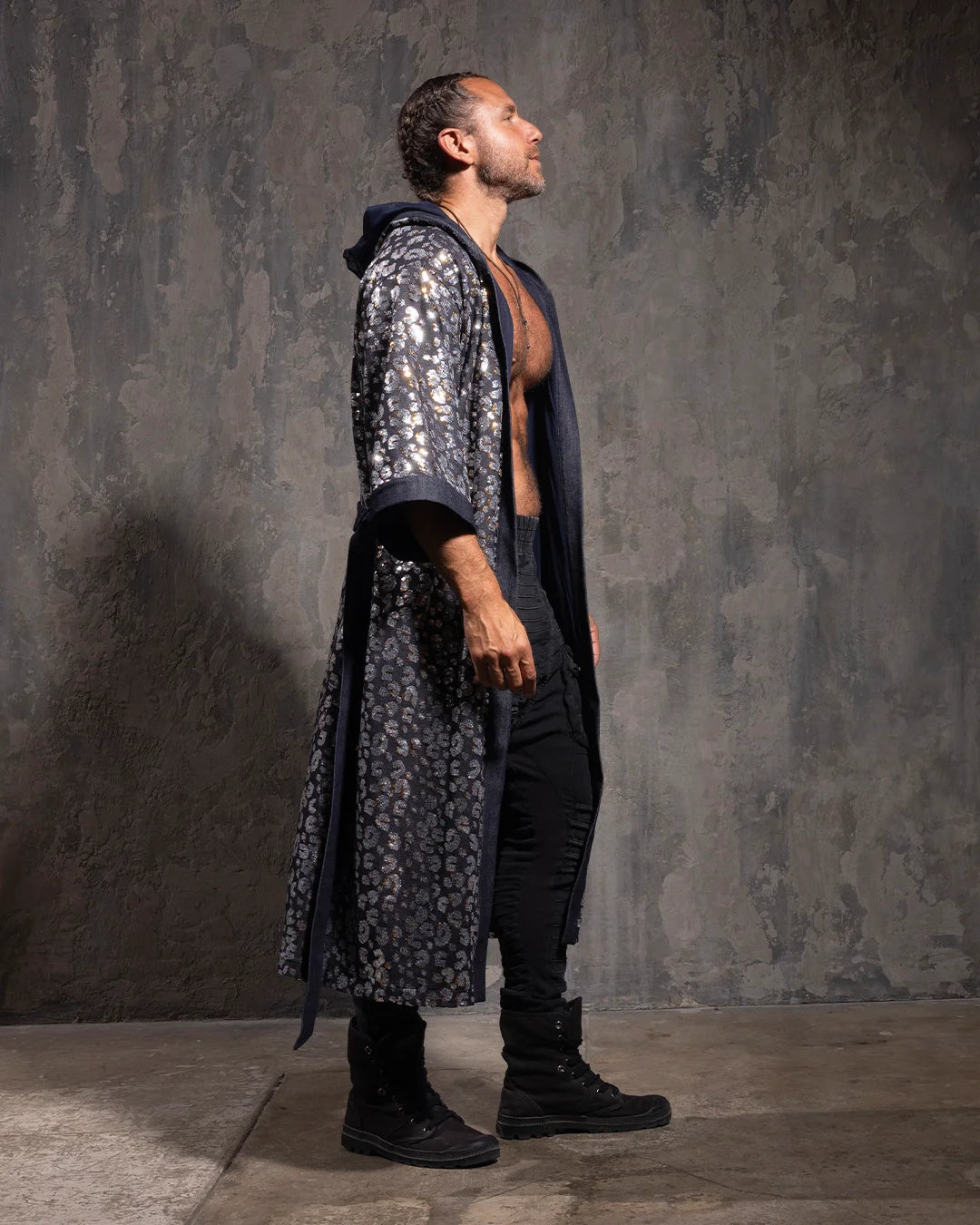 Man wearing a long, denim-colored hooded kimono with sequin leopard print details against a textured gray wall.