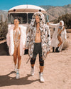 Bare-chested male model outdoors in the desert, wearing an open Hooded Wolverine eco-fur jacket with hood up, with female models and an Airstream