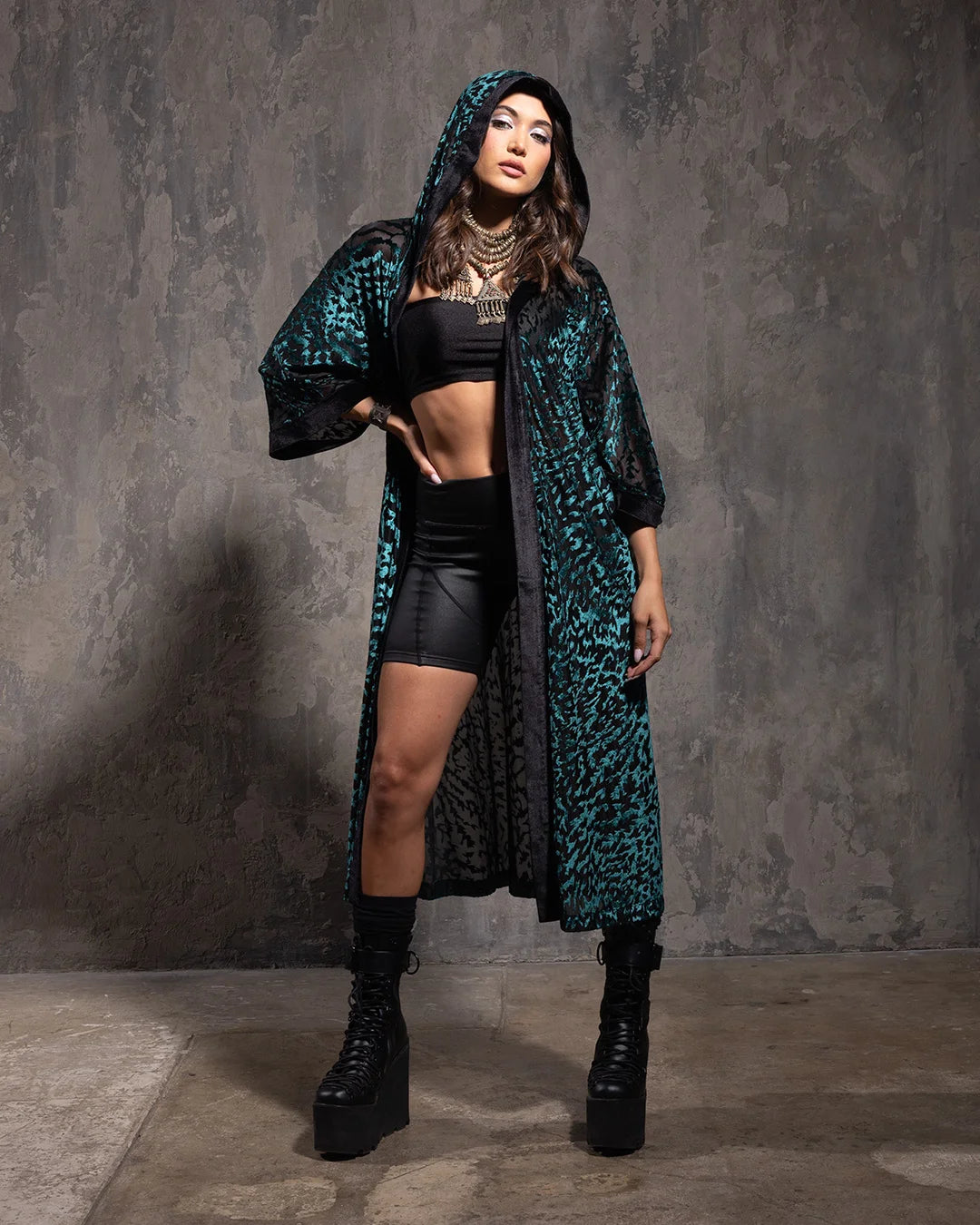 Woman wearing a long emerald green and black tiger print hooded kimono over a black outfit against a gray textured wall.