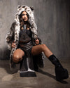 Woman wearing a fake fur coat with cheetah print, over sized hood with ears, over a black top, silver skirt, and black boots on a gray background.