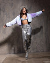 Woman wearing a futuristic outfit with SpiritHoods tie-dye purple and blue Mermaid Kitty Crop Jacket, silver pants, and boots against a gray background.