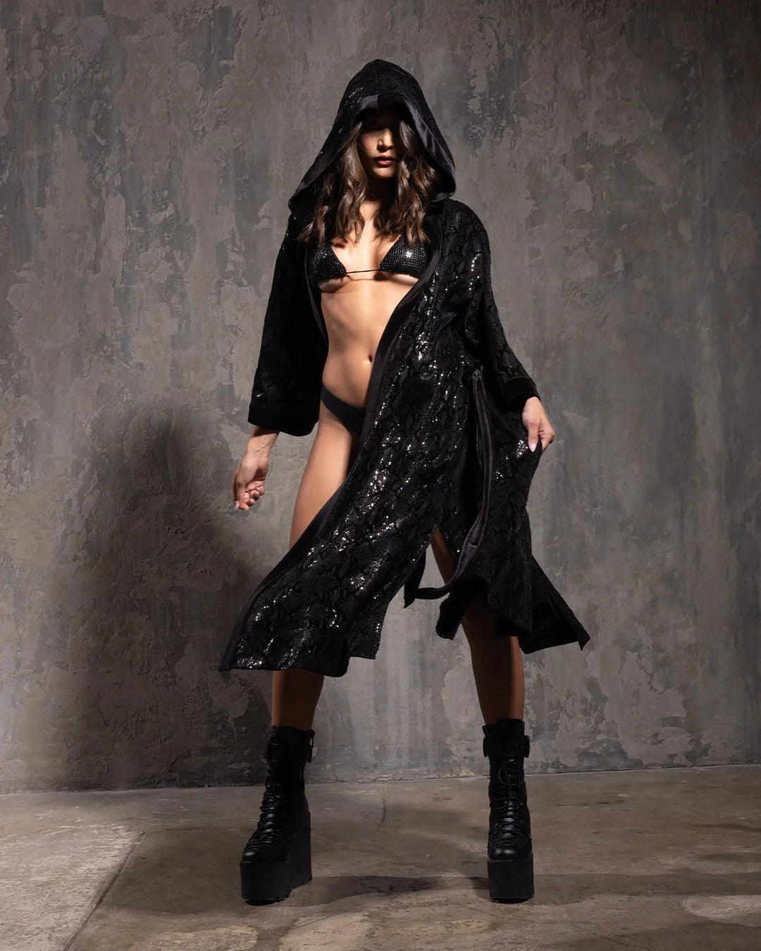 A Woman wearing a black sequin hooded festival kimono with a sparkling snakeskin print in front of a textured wall background
