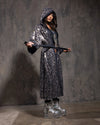 A Woman wearing a sparkling hooded kimono with silver and gold sequin leopard print and silver boots against a textured gray background