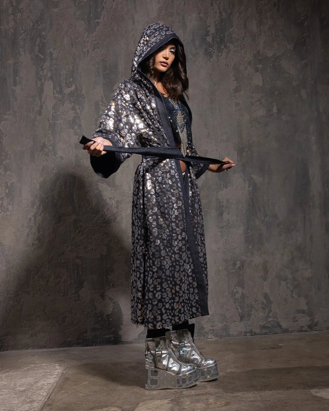 A Woman wearing a sparkling hooded kimono with silver and gold sequin leopard print and silver boots against a textured gray background
