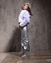 Woman wearing a light purple fake fur coat cropped at the waist with zipper closure and silver pants with a textured gray background