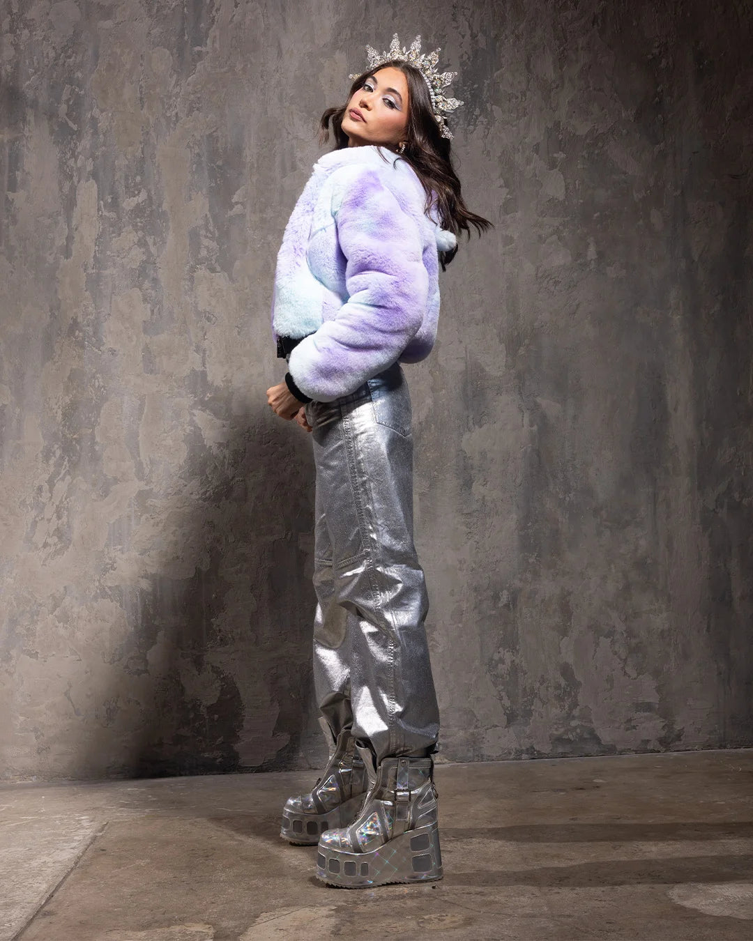 Woman wearing a light purple fake fur coat cropped at the waist with zipper closure and silver pants with a textured gray background