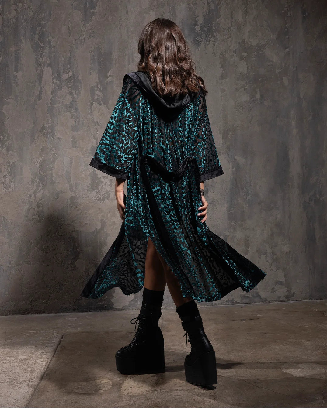 Woman wearing a dark green tiger print textured velvet kimono with a hood against a gray textured wall.