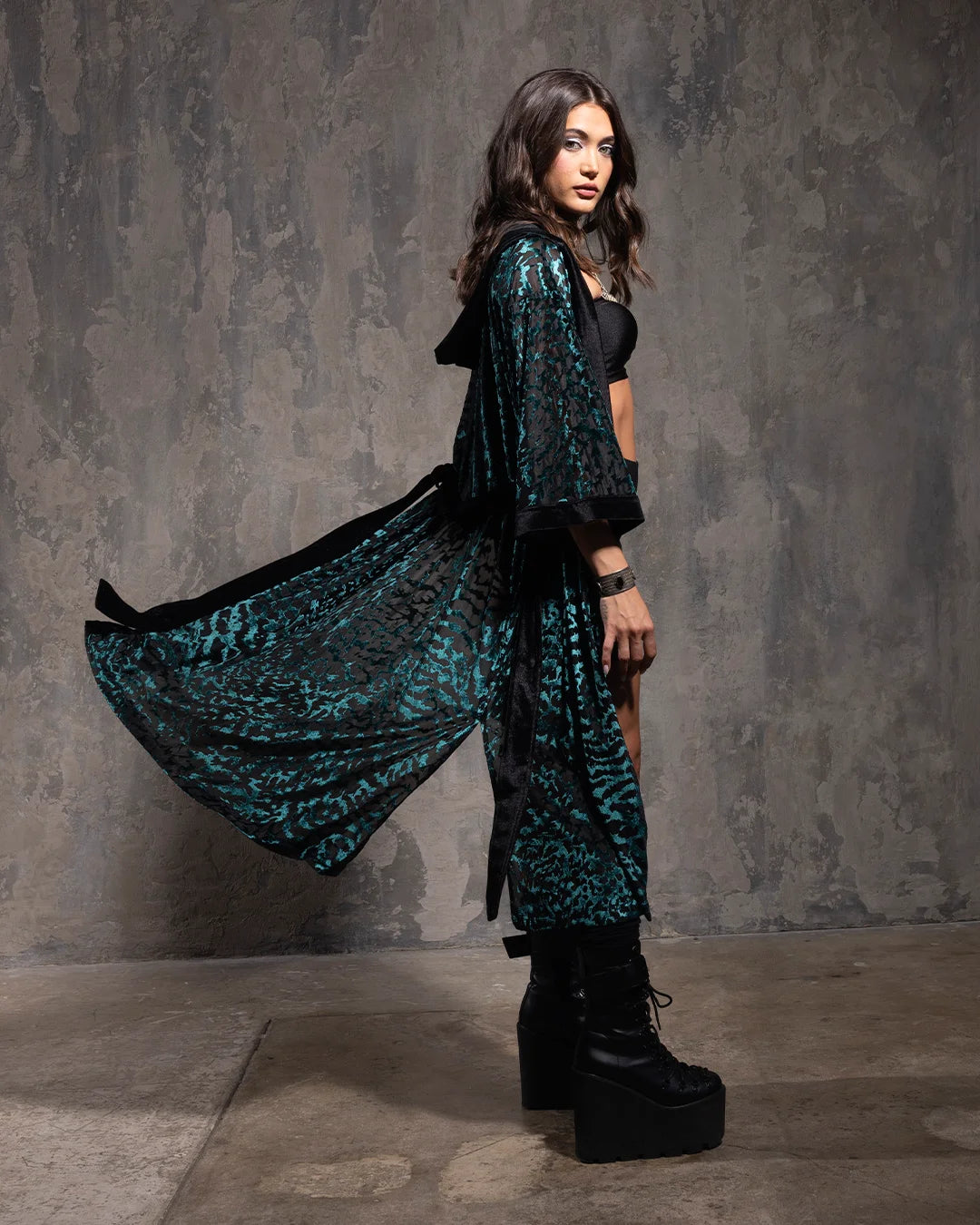 Woman wearing a long emerald green and black tiger print hooded kimono over a black outfit against a gray textured wall.