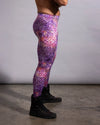 A side-profile view of a person wearing purple patterned leggings with a pink, orange, and white abstract print, paired with black boots, standing against a plain gray studio backdrop.