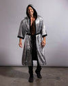 A man modeling a long, hooded silver sequin robe with black trim, worn open over a black shirt and black pants, standing against a plain gray studio backdrop.
