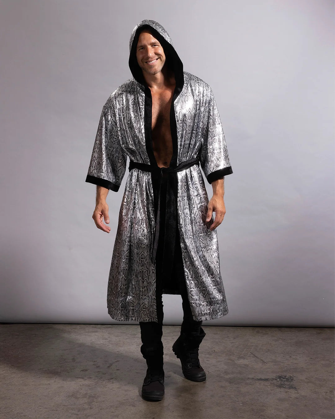 A man modeling a long, hooded silver sequin robe with black trim, worn open over a black shirt and black pants, standing against a plain gray studio backdrop.