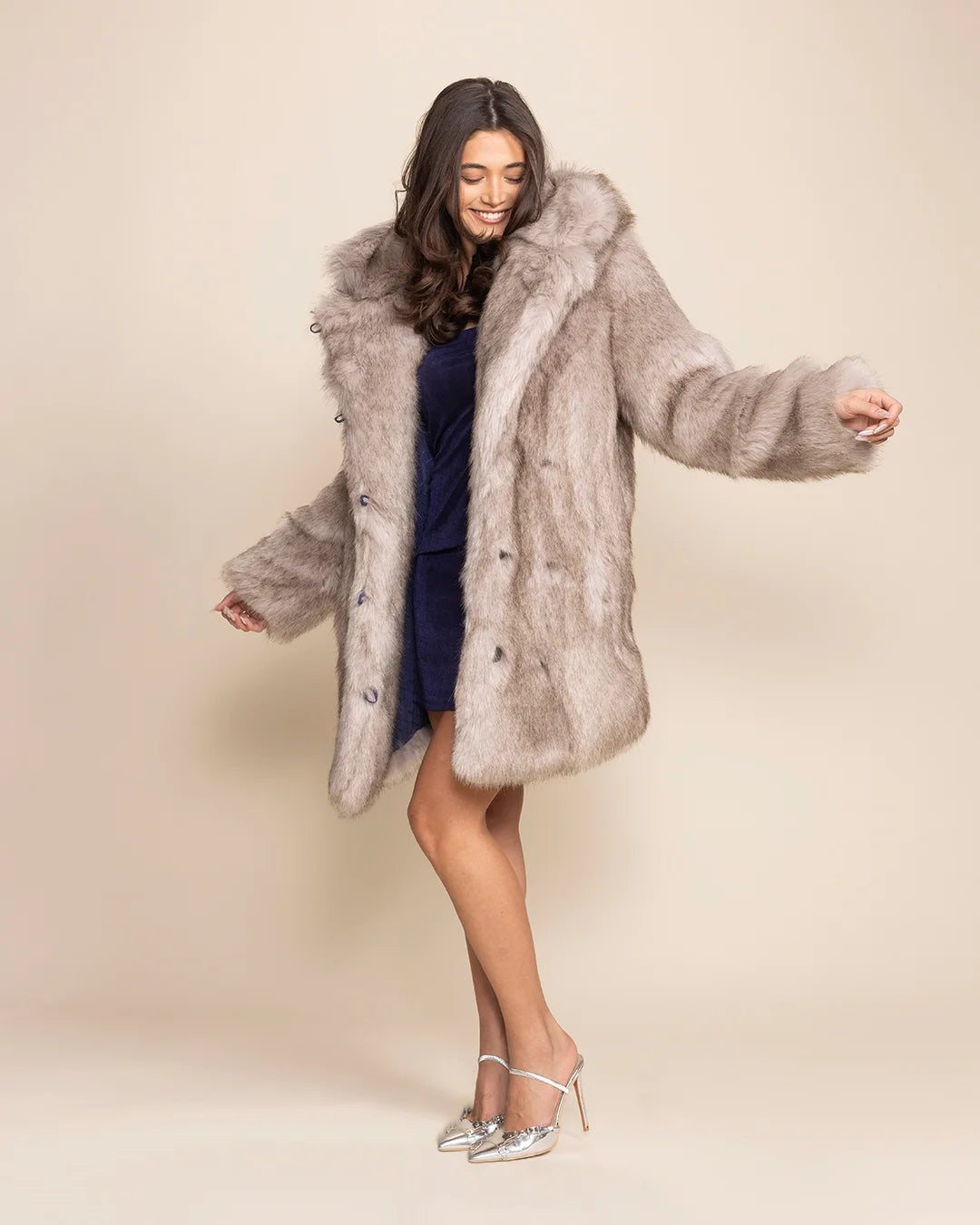 A Woman dancing in a heavyweight silvery grey faux fur winter coat with her arms out, looking happy.