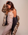 Woman wearing a SpiritHoods Classic leopard print fake fur coat over a beige top and striped pants against a plain background. Vegan Fashion Brand - Custom Faux Fur Coats and Jackets