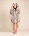 Woman wearing an extra plush silvery grey fake fur coat, with wolf ears showing a blue liner, holding the sides of the hood while she smiles. 100% Faux Fur Coats and Jackets