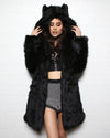 Dark haired girl looking down wearing realistic black wolf coat with hood and ears up, against a white background.