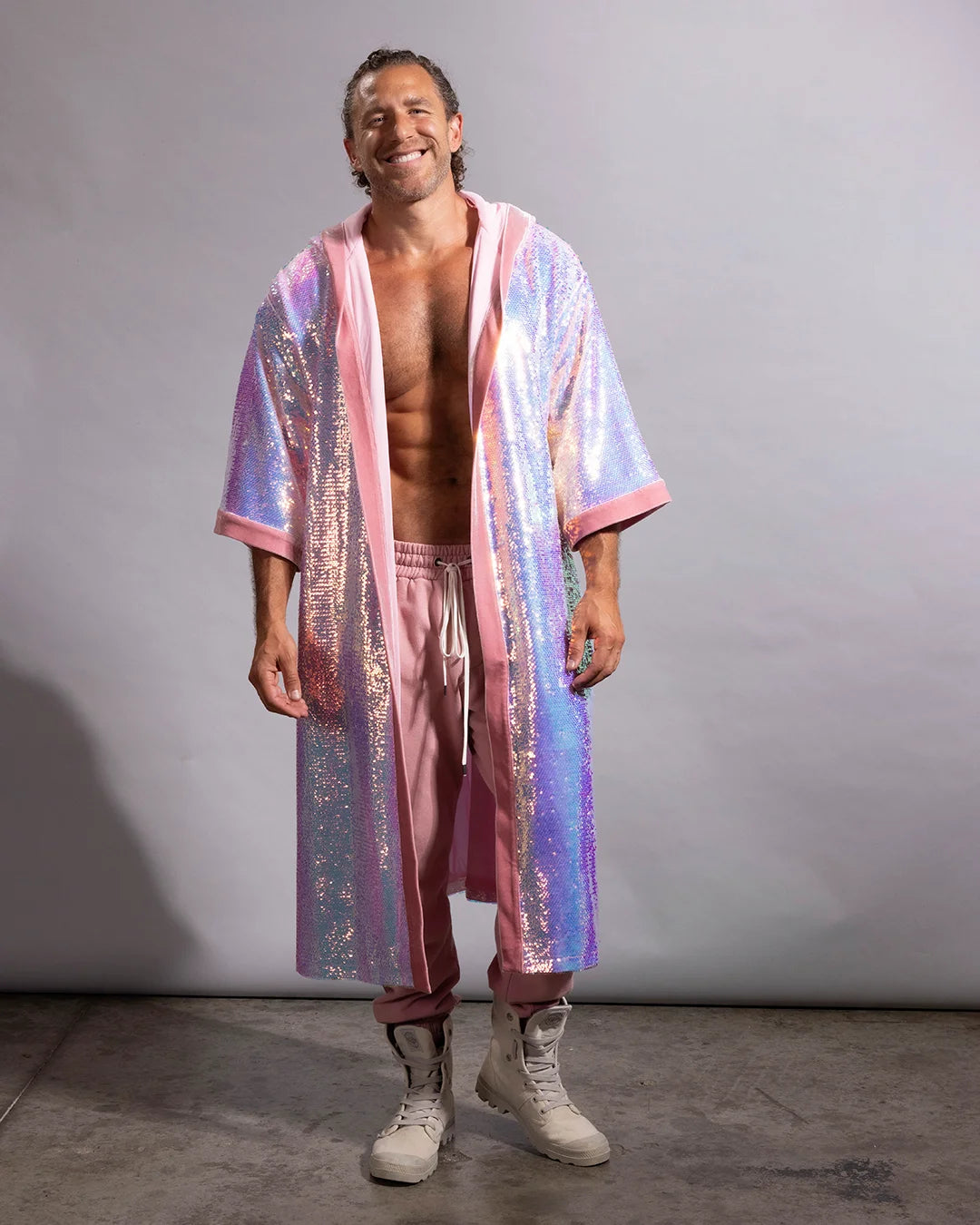 A man stands against a plain light gray studio backdrop wearing a sparkling iridescent pink kimono robe with pink trim, pink drawstring pants, and off-white lace-up boots.