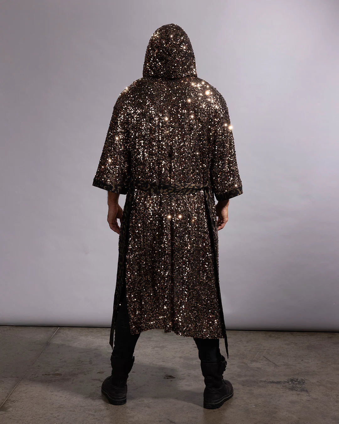 A person viewed from the back wearing a long, hooded kimono robe covered in dense metallic sequins, standing against a plain light gray studio backdrop.